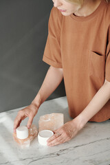 Close-up of young assistant of photographer putting two jars of facial cream on quartz platform on marble table before shooting