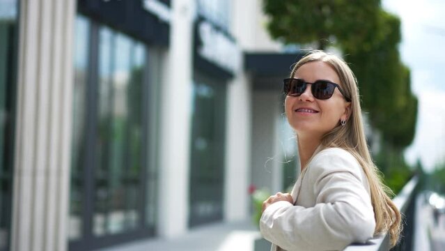 Happy young woman in sunglasses and white jacket in the street. Blonde takes off her glasses and smiles to the camera. Blurred backdrop.