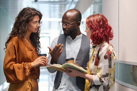 Two young businesswomen listening to explanation of confident African American male colleague while looking at him at meeting
