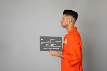 Mug shot of prisoner in orange uniform with board on grey background, side view