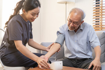 Obraz premium Pressure measurement of hypertension in elderly man sitting on couch, young woman nurse or caregiver care checking, measuring heart, blood by using sphygmomanometer monitor at home after retirement.