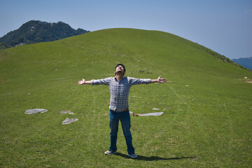 Fototapeta premium Happy guy in high altitude green grassland during day time arms outstretched