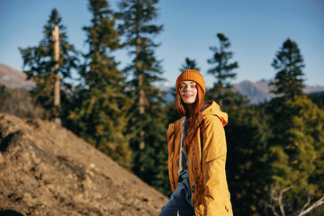 Woman hiking in the mountains in the autumn, a smile and happiness in a yellow cape with red hair full-length standing in front of trees and mountains in the sunset light