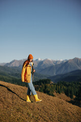 Woman autumn full-length walk on the hill smile with teeth and look at the mountains in a yellow raincoat and jeans happy hiking trip, freedom lifestyle 