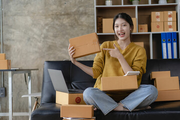 Asian woman working at home with yellow box and laptop for taking orders, sme business ideas on parcel delivery