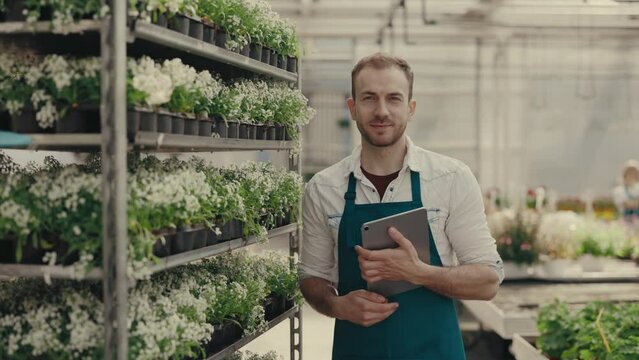 Man gardener is examining flowers holding the digital tablet and looking at the camera. Male greenhouse worker portrait, happy botanist, small business owner. People and professions concept.