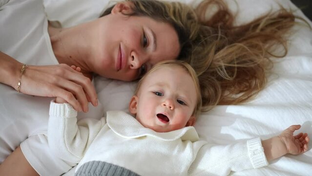 Curios little girl looking around and looking at camera as happy woman kissing child hand lying with baby on white soft cozy bed. High angle view portrait of Caucasian daughter and mother resting