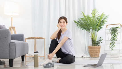 Beautiful Asian woman in sportswear is warming up or doing light exercises in the house with dumbbells and a protein shake or bottle of water and using a laptop at home, Healthy exercise.