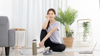 Beautiful Asian woman in sportswear is warming up or doing light exercises in the house with dumbbells and a protein shake or bottle of water and using a laptop at home, Healthy exercise.