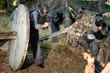 Team eliminating opponents on paintball field. People in protective outwear and helmets aiming with...