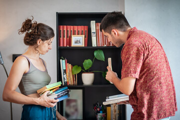 A relaxed young couple puts various kinds of books on a shelf in the room of their apartment