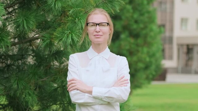 Middle-aged caucasian woman in glasses and white shirt looks at the camera. Female teacher in university garden. Beginning of the school year.