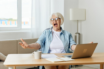 Fototapeta na wymiar an admiring, happy elderly woman is sitting at her desk working on a laptop and spreading her hand to the side shouts with happiness, rejoicing at success