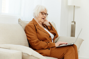 a thoughtful, wise, elderly woman writes an article on a laptop while sitting at home on the sofa in a comfortable environment in a beautiful brown suit and thoughtfully holds her hand near her face