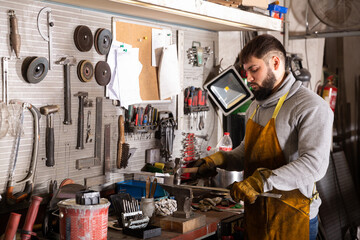Young bearded blacksmith shaping metal parts in metalworking workshop.. - Powered by Adobe