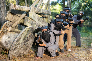 Fototapeta premium Tactical paintball team fighting on outdoor location. Men and women shooting with paintball markers, hiding behind barricade.
