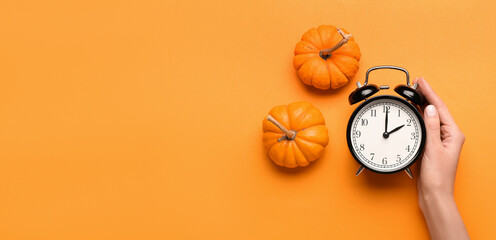 Female hand with alarm clock and pumpkins on orange background with space for text