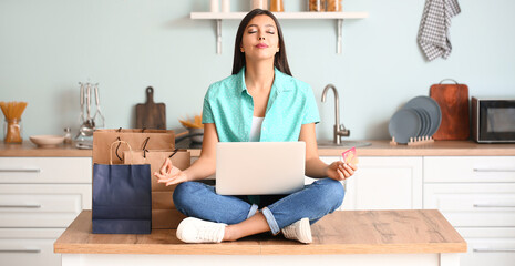 Meditating woman with laptop, credit card and shopping bags in kitchen