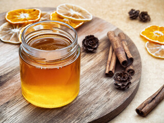 Transparent jar with honey on a wooden round tray, surrounded by cinnamon, dried orange and lemon in warm light colors.