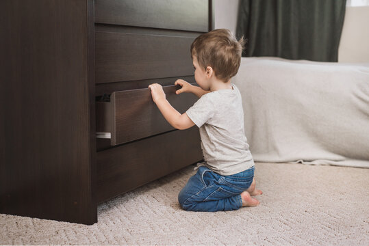 curious two-year-old boy in gray T-shirt and jeans plays at home and opens chest of drawers. Child safety latches on wardrobe. Games without parental supervision. Safe children's space. Selected focus