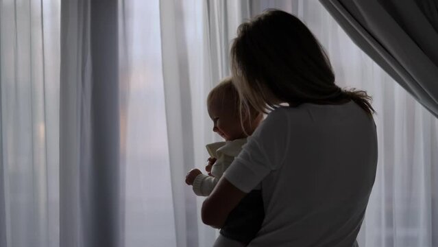 Silhouette of mother kissing baby girl in slow motion standing at window indoors. Back view medium shot of happy loving Caucasian woman enjoying morning with infant daughter