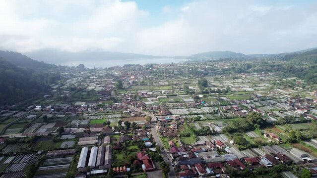 Many greenhouses and strawberry plantations at village in old crater, aerial view of Pancasari, Bratan lake seen at distance. Panoramic shot from height, light clouds hang low in the air