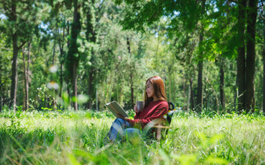 Naklejka na ściany i meble Portrait of a beautiful young asian woman reading a book and drinking coffee while sitting on a camping chair in the park