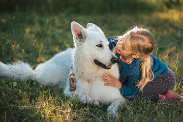 Cute blonde Caucasian little girl sitting on the grass and caressing her pet dog, a beautiful White...