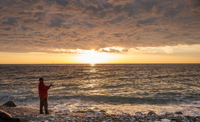 Fototapeta premium December 6, 2021, Sochi, Russia. An elderly man fishing on the embankment in Sochi against the backdrop of the setting sun and amazing clouds