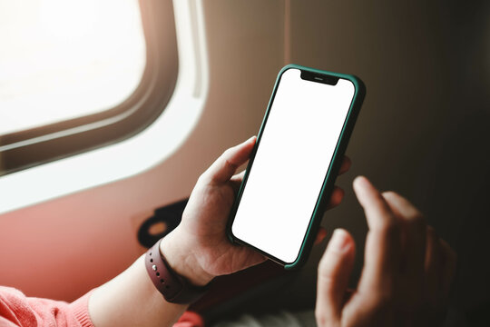 Mockup image of a hand holding a black mobile phone with blank desktop screen next to an airplane window