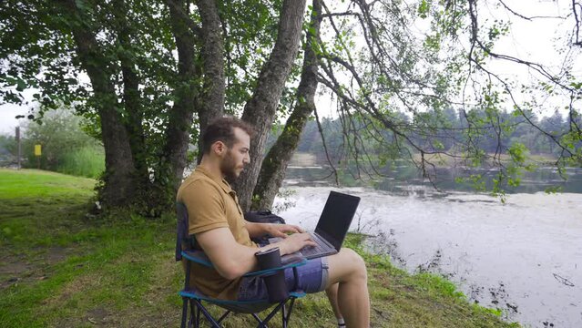 Working with a computer in nature.
Young man sitting in camping chair working with his laptop.

