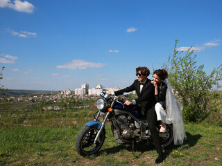 Fototapeta na wymiar Unusial wedding on the motocycle. the bride in a leather jacket, the groom in glasses. Happy couple smiling outdoors