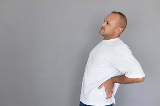 Portrait of mature Caucasian man touching aching low back. Balding guy wearing white T-shirt and jeans stretching his back. Low back pain concept