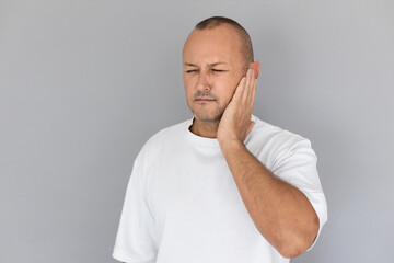 Portrait of mature Caucasian man suffering from earache. Balding guy wearing white T-shirt touching his ear against gray background. Health problems concept