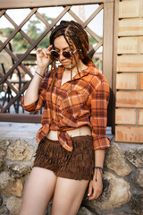 a girl with a dreadlocked hairstyle poses in summer outdoor, bright sunlight, dressed in a plaid shirt and shorts