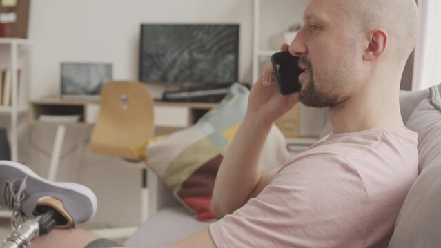Tilt down slowmo of Caucasian man with prosthetic leg talking on mobile phone sitting on sofa at home
