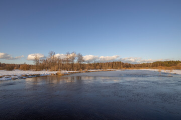March sunny evening by the river. Blue sky over the horizon. A picturesque landscape, early spring, a river with snow-covered banks, dry grass and bushes. The first thaws, the snow is melting