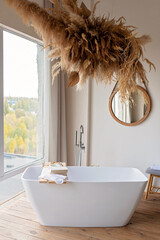 Comfortable bright bathroom with a boho-chic interior design, a free-standing white bath against the background of a panoramic window. hanging decor from dry grass. Soft selective focus.