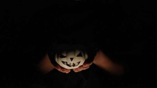 Hands from the darkness holding a white pumpkin for Halloween, light from the fire.