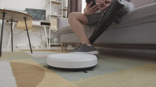 Cropped slowmo of unrecognizable man with prosthetic leg connecting robotic vacuum cleaner to smartphone, sitting on sofa in living room