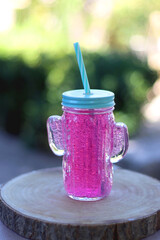 Cactus shaped glass filled with pink soda in the garden. Refreshing summer drink. Selective focus.