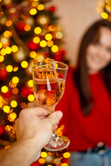 Two glasses of champagne in hands of a man and young woman, close up. Blurred girl, christmas tree and warm lights on the background