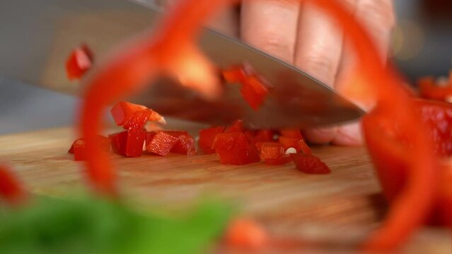 Camera movement on hands cut red bell pepper on wooden cutting board on background of greens in kitchen. Chop vegetables. Cooking food.