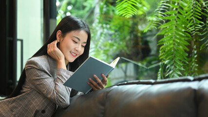 Attractive millennial woman reading book on couch in beautiful day. Leisure activity concept