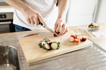 Obraz na płótnie Canvas A man wearing a white t-shirt chopping up strawberries in a kitchen on a wooden board with a knife