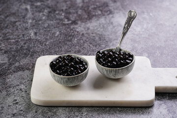 Fototapeta na wymiar Cooked tapioca pearls for trendy bubble boba ice tea in two small grey ceramic bowls on marble board on dark grey concrete background