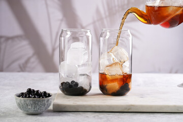 Female hand pouring black tea into transparent glass with ice cubes and cooked tapioca pearls for trendy bubble boba ice tea, two small grey ceramic bowls on marble board on grey concrete background