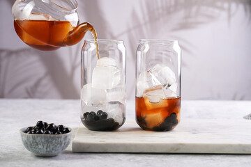 Female hand pouring black tea into transparent glass with ice cubes and cooked tapioca pearls for trendy bubble boba ice tea, two small grey ceramic bowls on marble board on grey concrete background