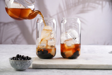 Female hand pouring black tea into transparent glass with ice cubes and cooked tapioca pearls for trendy bubble boba ice tea, two small grey ceramic bowls on marble board on grey concrete background