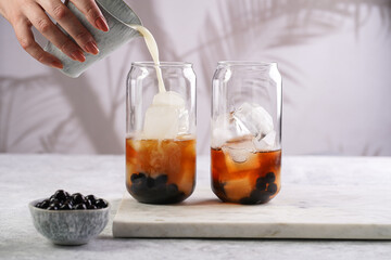 Female hand pouring milk into glass with black tea, ice cubes and cooked tapioca pearls for trendy bubble boba ice tea, two small grey ceramic bowls on marble board on grey concrete background